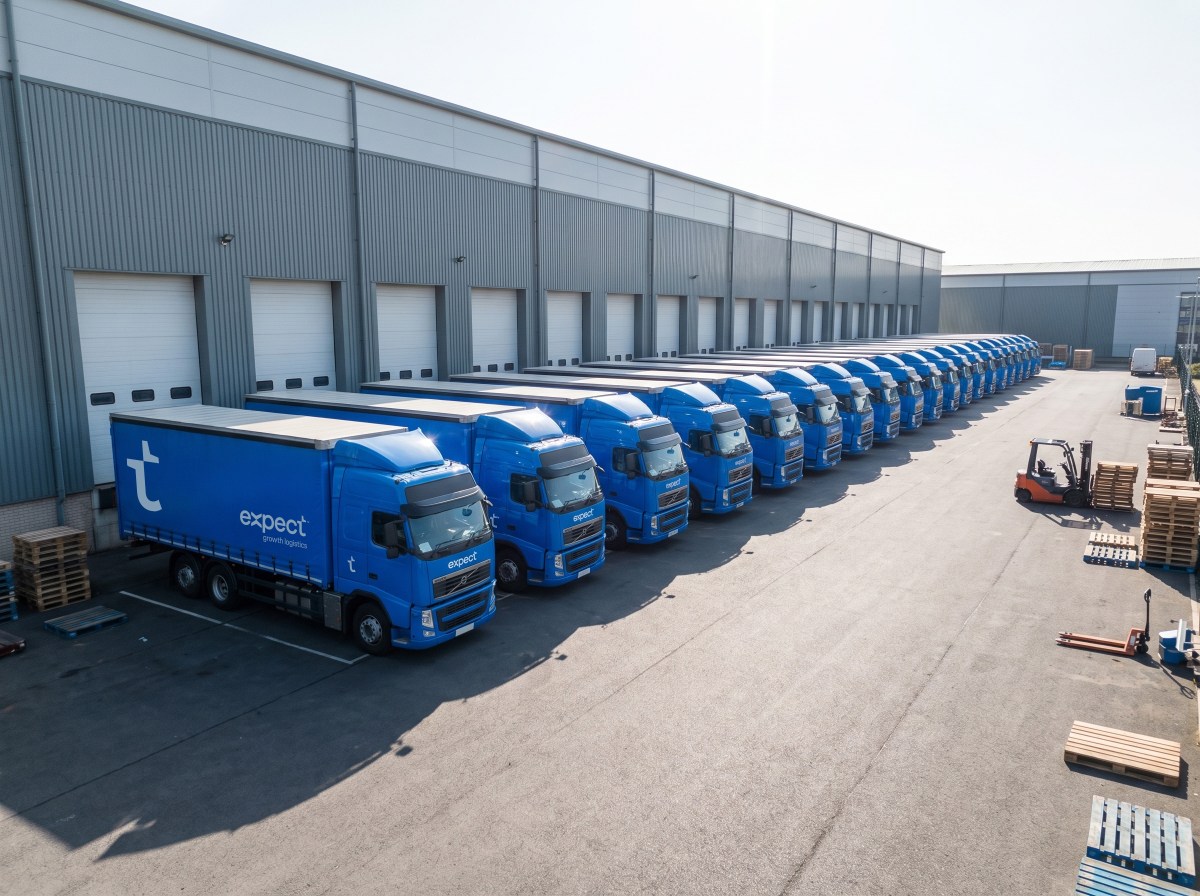Fleet of blue Expect Logistics lorries parked at loading bays outside a large distribution warehouse, with a forklift moving pallets nearby.
